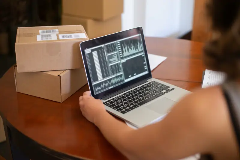 Person working on a laptop beside open shipping boxes. The screen shows charts.
