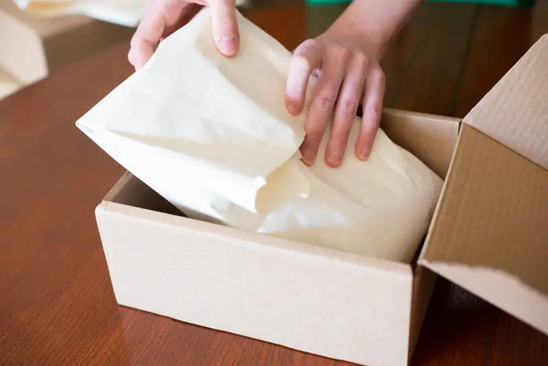 Hands wrapping an item in tissue paper inside a shipping box.