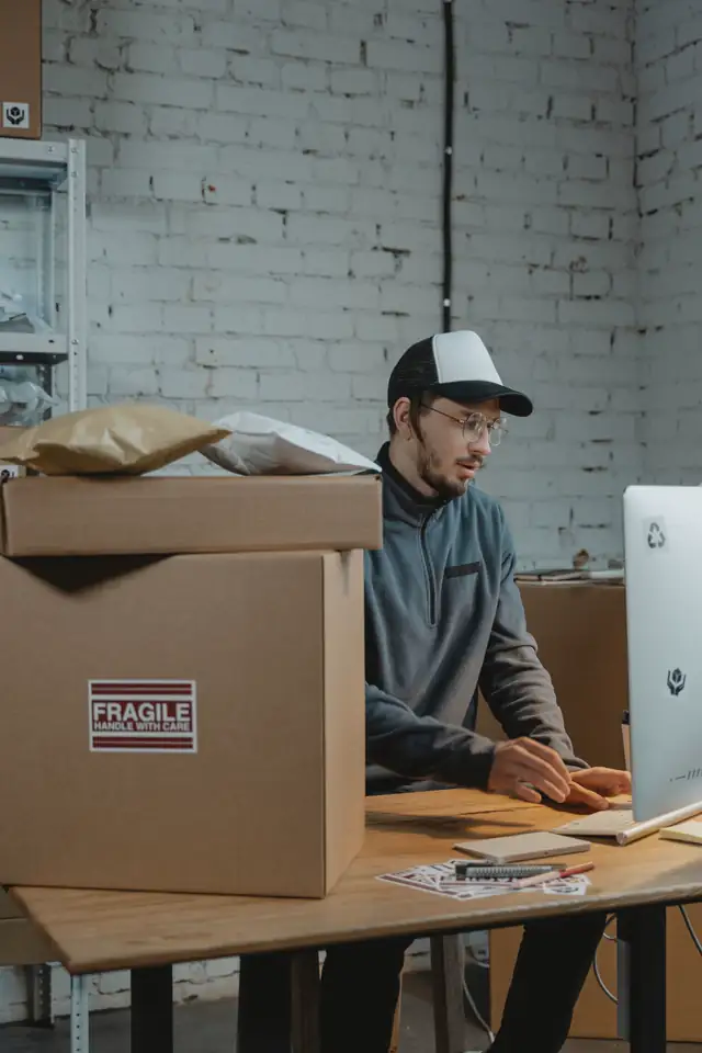 A person at a desktop computer, with shelves of shipping cartons behind them.